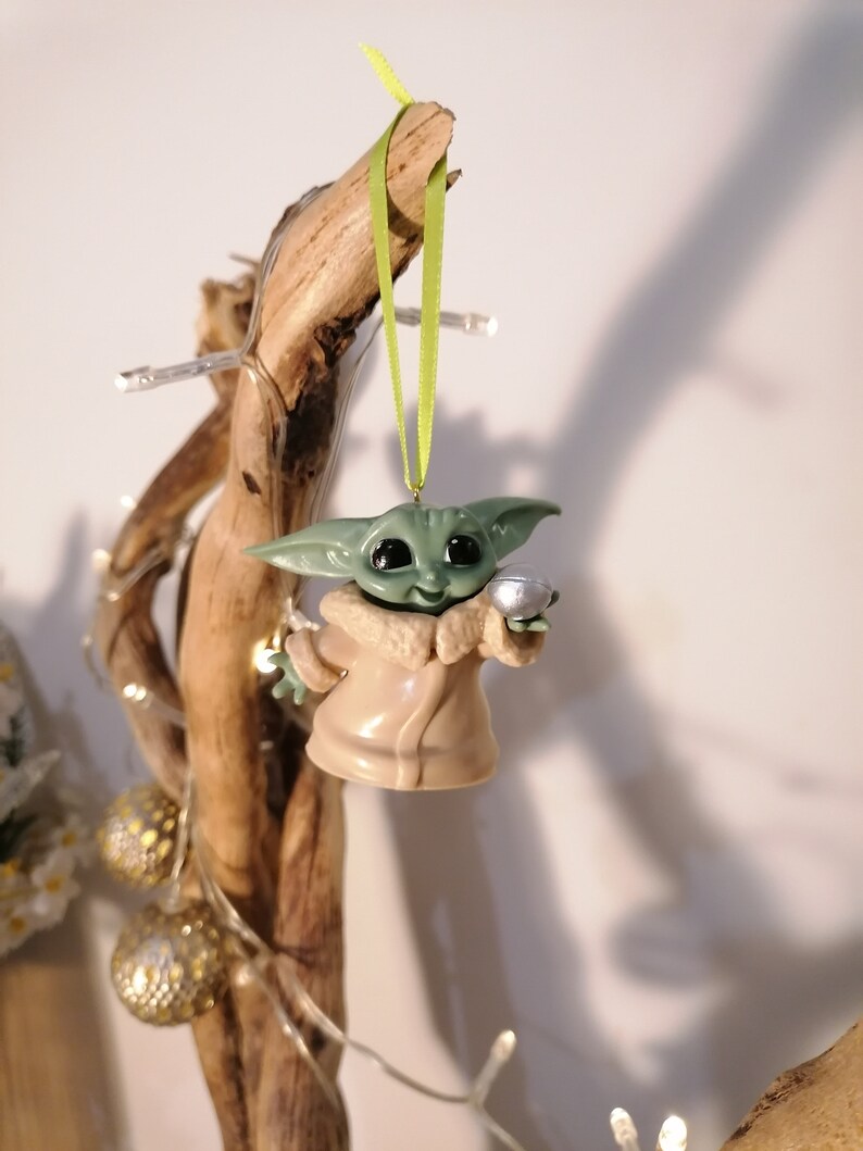 May include: A green and brown Christmas ornament featuring a character from the Star Wars franchise, holding a silver bell. The ornament is hanging from a green ribbon on a branch.