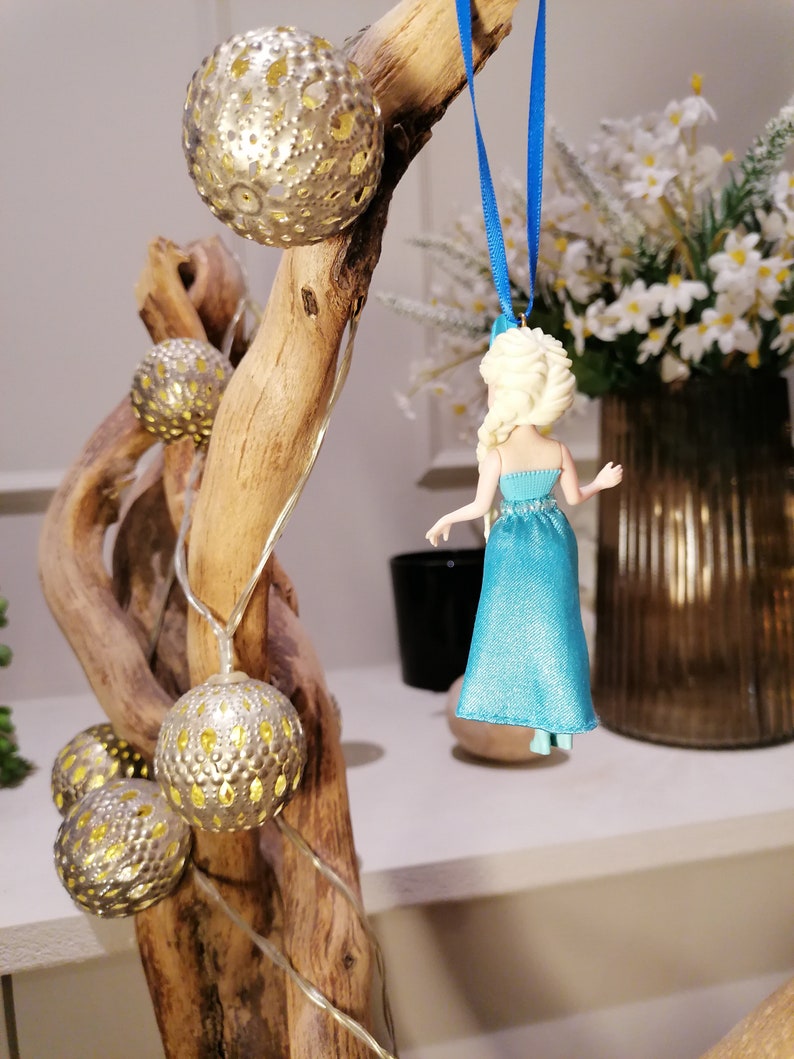 K&ouml;nnte beinhalten: Eine blaue und wei&szlig;e Figur einer Frau mit langen blonden Haaren, die an einem blauen Band an einem Treibholzast mit goldenen Lichterketten h&auml;ngt.