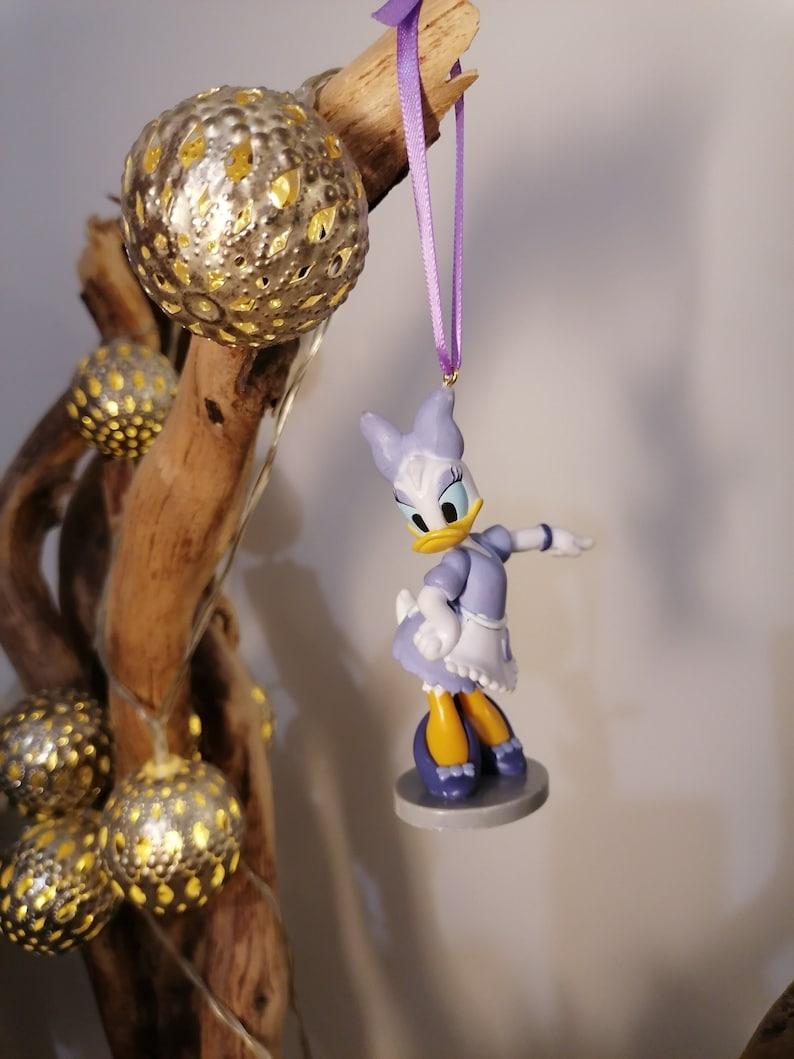 Puede incluir: Una figurita de pl&aacute;stico de un pato de dibujos animados que lleva un vestido azul y un delantal blanco. La figurita est&aacute; colgada de una cinta morada.