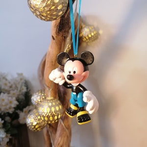 May include: A blue and black Mickey Mouse figurine ornament hanging from a blue ribbon on a wooden branch. The ornament is made of plastic and has a cartoonish style.