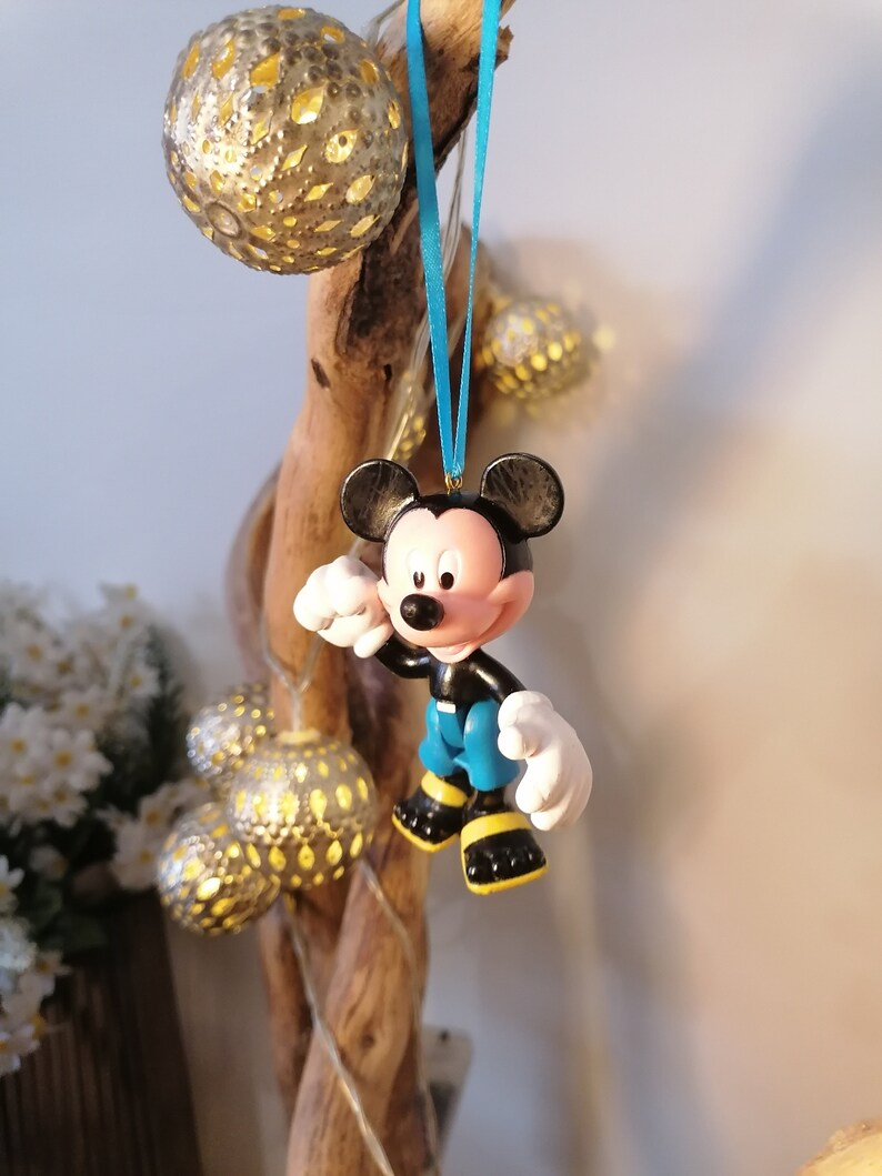 May include: A blue and black Mickey Mouse figurine ornament hanging from a blue ribbon on a wooden branch. The ornament is made of plastic and has a cartoonish style.