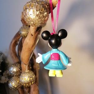 May include: A blue and pink plastic ornament of a cartoon character wearing a blue kimono with a pink sash. The character has black ears and a black face. The ornament is hanging from a pink ribbon.