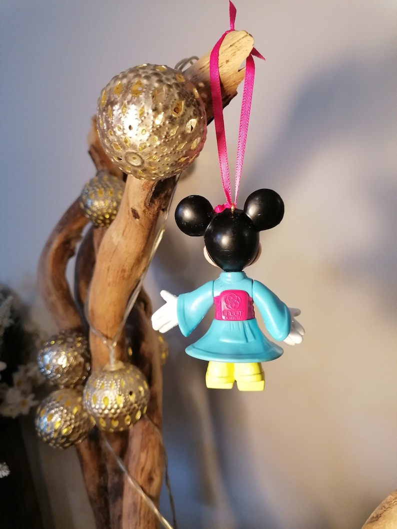 May include: A blue and pink plastic ornament of a cartoon character wearing a blue kimono with a pink sash. The character has black ears and a black face. The ornament is hanging from a pink ribbon.