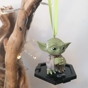 Puede incluir: Un adorno de Yoda verde colgando de una cinta verde. La figura sostiene un bast&oacute;n marr&oacute;n y est&aacute; de pie sobre una base hexagonal negra con las palabras "Star Wars" impresas en ella.