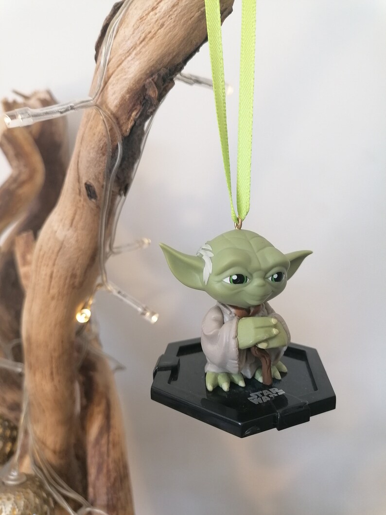 Puede incluir: Un adorno de Yoda verde colgando de una cinta verde. La figura sostiene un bast&oacute;n marr&oacute;n y est&aacute; de pie sobre una base hexagonal negra con las palabras "Star Wars" impresas en ella.