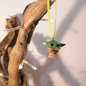 May include: A green and brown Yoda ornament hanging from a branch with a green ribbon. The ornament is a small, detailed figure of Yoda holding a cup.