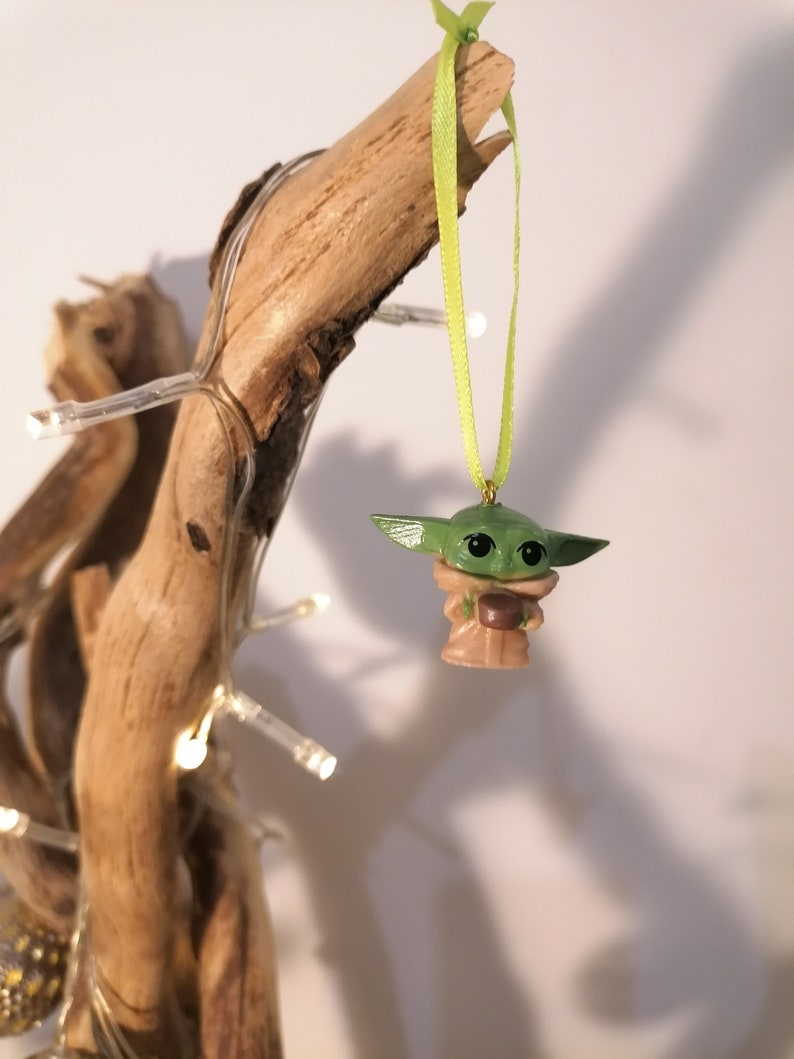May include: A green and brown Yoda ornament hanging from a branch with a green ribbon. The ornament is a small, detailed figure of Yoda holding a cup.