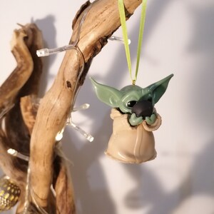 May include: A green and brown Yoda ornament hanging from a branch with a green ribbon. The ornament is holding a small, round object in its hands.
