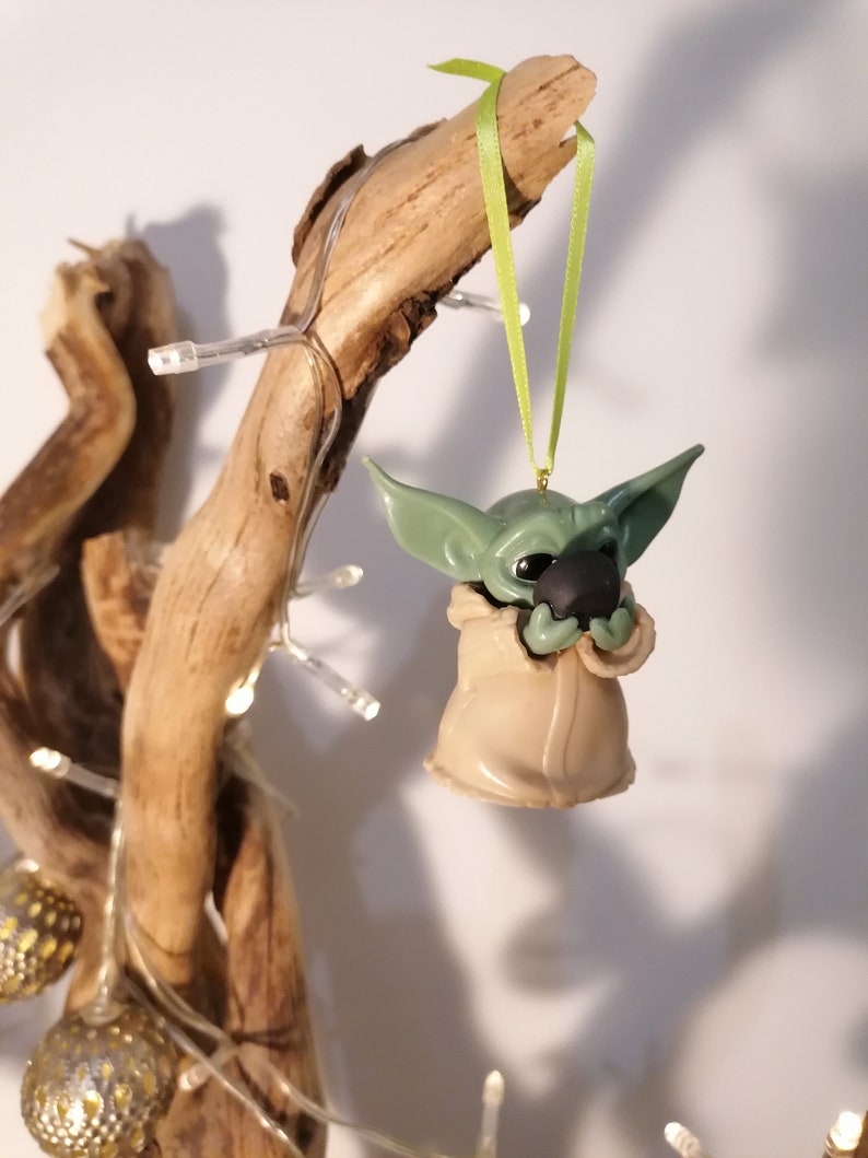 May include: A green and brown Yoda ornament hanging from a branch with a green ribbon. The ornament is holding a small, round object in its hands.