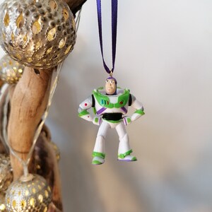 Puede incluir: Un adorno de plástico verde, blanco y morado de Buzz Lightyear de la franquicia Toy Story. El adorno está colgado de una cinta morada.