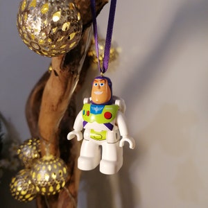 May include: A white plastic toy figurine of Buzz Lightyear from the Toy Story franchise, hanging from a purple ribbon. The figurine is wearing a green and purple spacesuit with a red button and a white wing emblem. The figurine is smiling and has a light brown face.