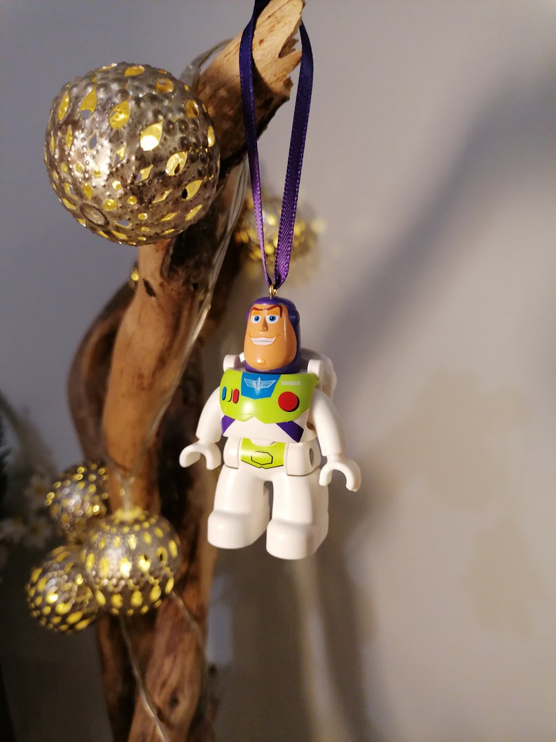 May include: A white plastic toy figurine of Buzz Lightyear from the Toy Story franchise, hanging from a purple ribbon. The figurine is wearing a green and purple spacesuit with a red button and a white wing emblem. The figurine is smiling and has a light brown face.