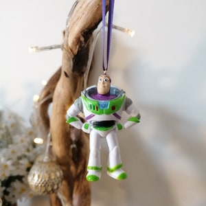 May include: A green, white, and purple plastic ornament of Buzz Lightyear from the Toy Story movie franchise. The ornament is hanging from a purple ribbon.