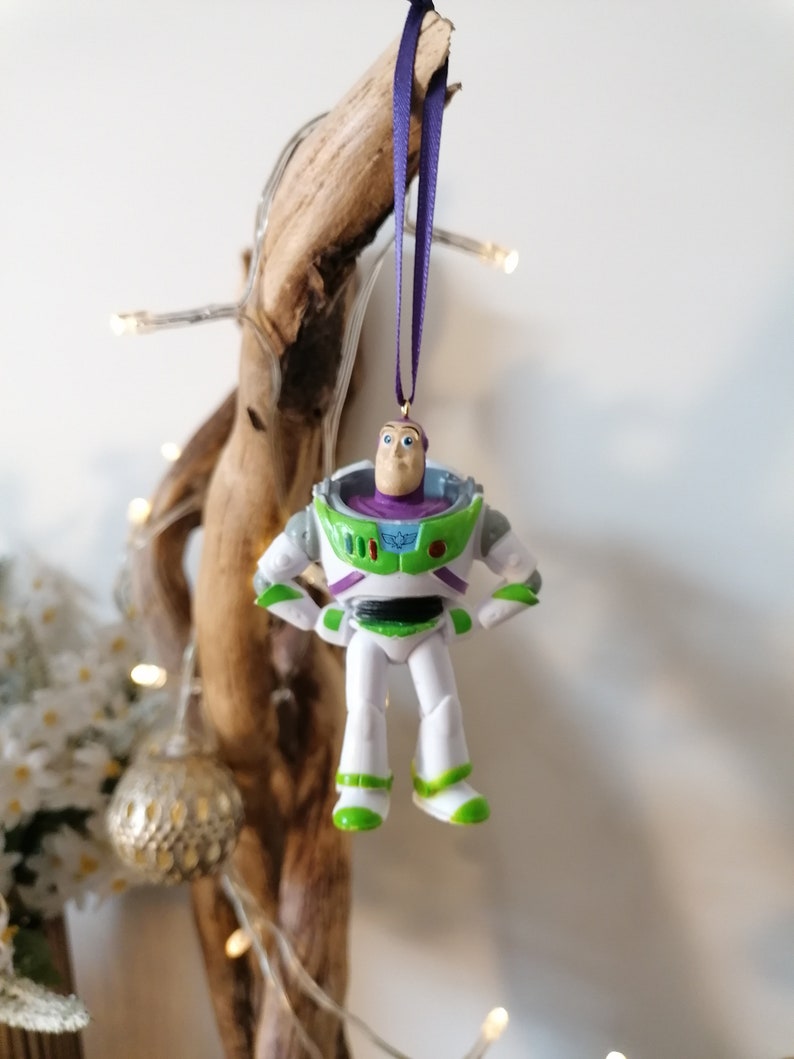 May include: A green, white, and purple plastic ornament of Buzz Lightyear from the Toy Story movie franchise. The ornament is hanging from a purple ribbon.