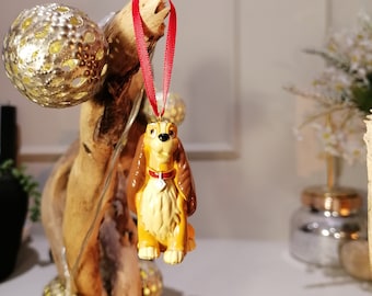 May include: A ceramic ornament of Lady, the dog from Disney's Lady and the Tramp, hanging from a red ribbon. The ornament is yellow and brown with a red collar and a tag.