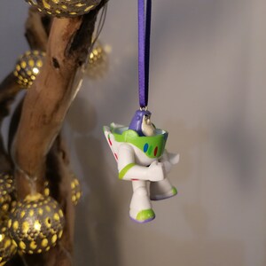 May include: A white and green Buzz Lightyear Christmas ornament with a purple ribbon hanging from a branch.