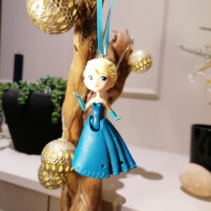 Peut inclure: Une figurine de princesse en plastique bleu, portant une robe bleue avec un ruban blanc autour du cou. La poup&eacute;e est suspendue &agrave; un ruban bleu sur une branche de bois.