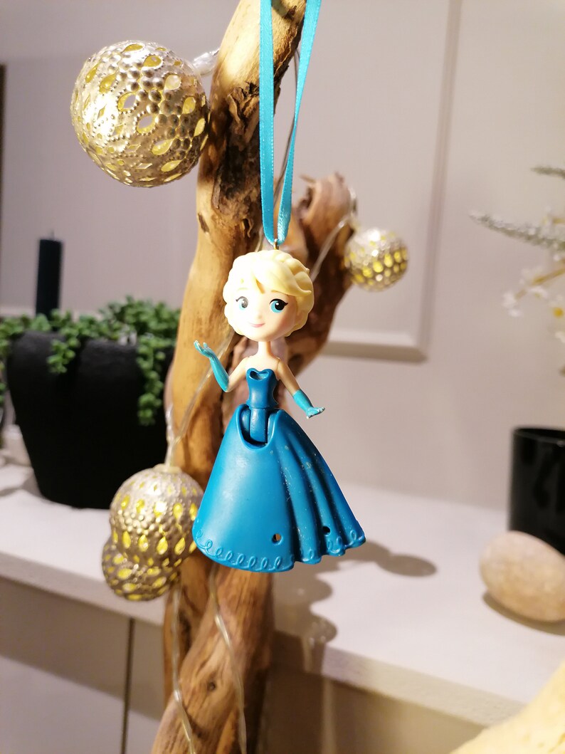 Peut inclure: Une figurine de princesse en plastique bleu, portant une robe bleue avec un ruban blanc autour du cou. La poup&eacute;e est suspendue &agrave; un ruban bleu sur une branche de bois.