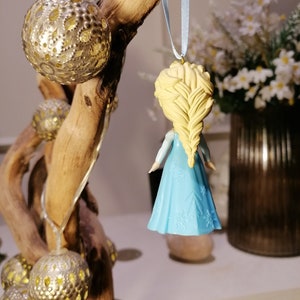 May include: A blue and white ornament of a girl with long blonde hair, wearing a blue dress with snowflake designs. The ornament is hanging from a blue ribbon.