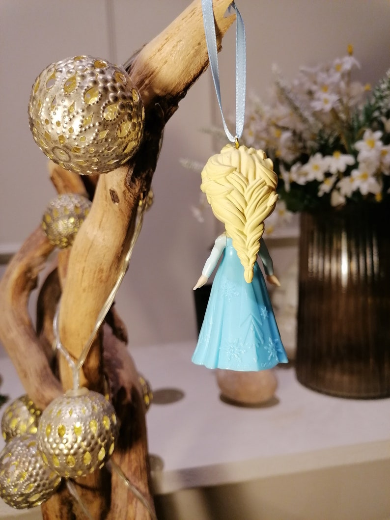 May include: A blue and white ornament of a girl with long blonde hair, wearing a blue dress with snowflake designs. The ornament is hanging from a blue ribbon.