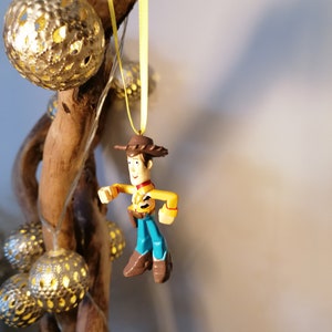May include: A Woody action figure ornament, featuring a yellow shirt, blue pants, and a brown cowboy hat and boots. The ornament hangs from a yellow ribbon. Decorative gold-colored sphere lights and a wooden branch are in the background.
