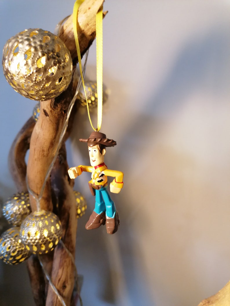 May include: A Woody action figure ornament, featuring a yellow shirt, blue pants, and a brown cowboy hat and boots. The ornament hangs from a yellow ribbon. Decorative gold-colored sphere lights and a wooden branch are in the background.