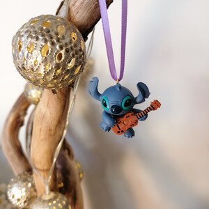 Op de afbeelding: Blauw Stitch-personage ornament dat een bruine gitaar bespeelt, hangend aan een paars lint aan een houten tak met zilveren decoratieve ballen.