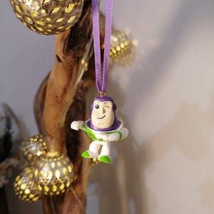 May include: Purple and white plastic ornament of Buzz Lightyear from the Toy Story franchise, hanging from a purple ribbon on a wooden branch.