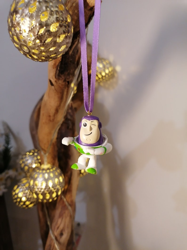 May include: Purple and white plastic ornament of Buzz Lightyear from the Toy Story franchise, hanging from a purple ribbon on a wooden branch.