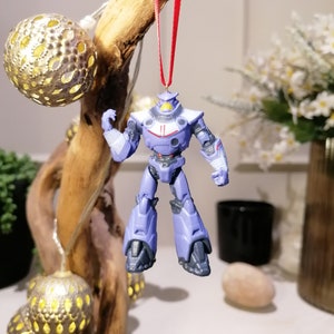 Puede incluir: Un adorno de Navidad de robot morado y azul que cuelga de una cinta roja. El robot tiene una franja blanca en el pecho y una visera negra.