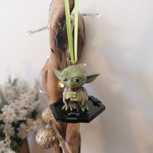 Puede incluir: Un adorno de Yoda verde colgando de una rama con una cinta verde. El adorno est&aacute; sobre una base hexagonal negra con el logotipo de Star Wars.