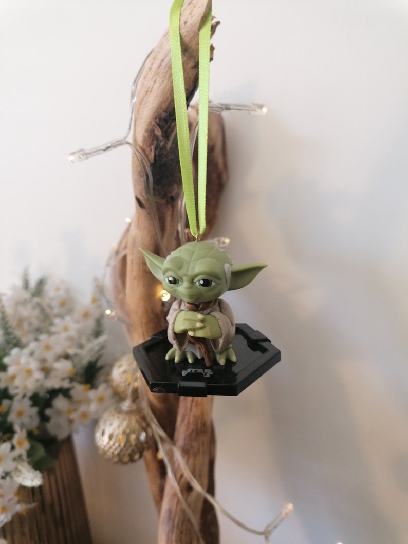 Puede incluir: Un adorno de Yoda verde colgando de una rama con una cinta verde. El adorno est&aacute; sobre una base hexagonal negra con el logotipo de Star Wars.