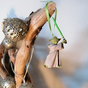 Puede incluir: Un adorno navide&ntilde;o de Yoda con un sable de luz verde cuelga de una rama de madera. La figura de Yoda lleva una t&uacute;nica beige. Adornos esf&eacute;ricos plateados y luces de cadena tambi&eacute;n est&aacute;n unidos a la rama.