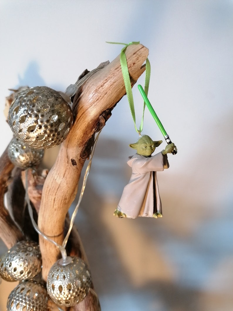 Puede incluir: Un adorno navide&ntilde;o de Yoda con un sable de luz verde cuelga de una rama de madera. La figura de Yoda lleva una t&uacute;nica beige. Adornos esf&eacute;ricos plateados y luces de cadena tambi&eacute;n est&aacute;n unidos a la rama.