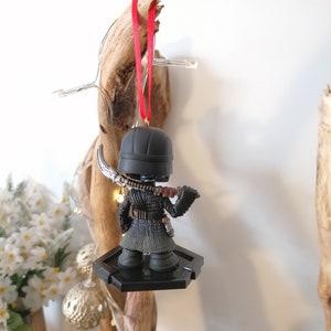 May include: Black and red Christmas ornament featuring a small figurine of a character from the Star Wars franchise. The character is wearing a black hooded cloak and holding a lightsaber. The ornament is hanging from a red ribbon.