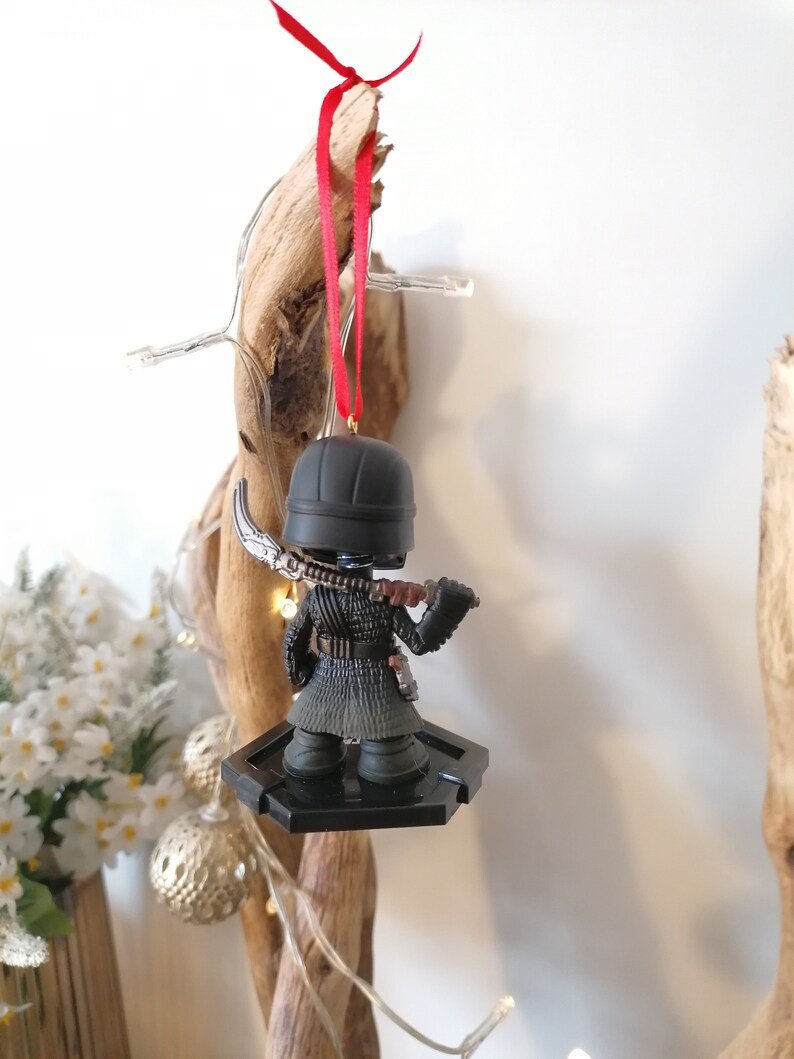 May include: Black and red Christmas ornament featuring a small figurine of a character from the Star Wars franchise. The character is wearing a black hooded cloak and holding a lightsaber. The ornament is hanging from a red ribbon.