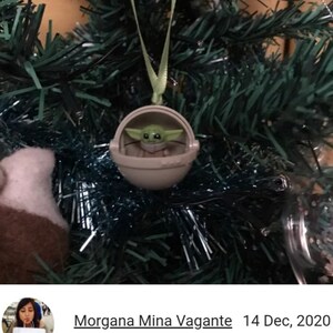 May include: A Christmas tree ornament featuring a small, green-skinned character in a tan pod. The ornament is suspended by a green ribbon against a backdrop of green tree branches and tinsel. The character is from the Star Wars franchise.