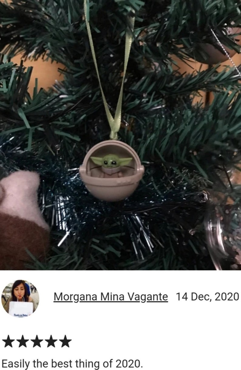 May include: A Christmas tree ornament featuring a small, green-skinned character in a tan pod. The ornament is suspended by a green ribbon against a backdrop of green tree branches and tinsel. The character is from the Star Wars franchise.