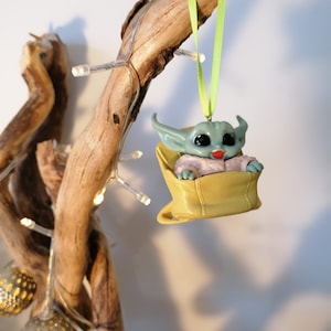 May include: A green and blue character ornament hanging from a branch with a green ribbon. The character is sitting in a yellow bag with a white lining. The ornament is a popular character from the Star Wars franchise.