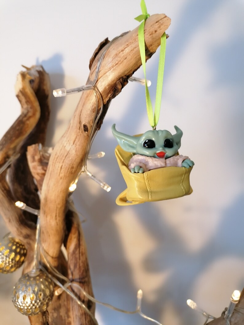 May include: A green and blue character ornament hanging from a branch with a green ribbon. The character is sitting in a yellow bag with a white lining. The ornament is a popular character from the Star Wars franchise.