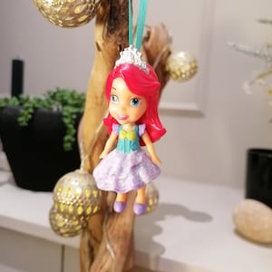 Peut inclure: Une petite poup&eacute;e en plastique aux cheveux roux, v&ecirc;tue d'une robe violette &agrave; paillettes, d'un haut bleu et jaune et d'une couronne argent&eacute;e. La poup&eacute;e est suspendue &agrave; un ruban turquoise sur une branche.