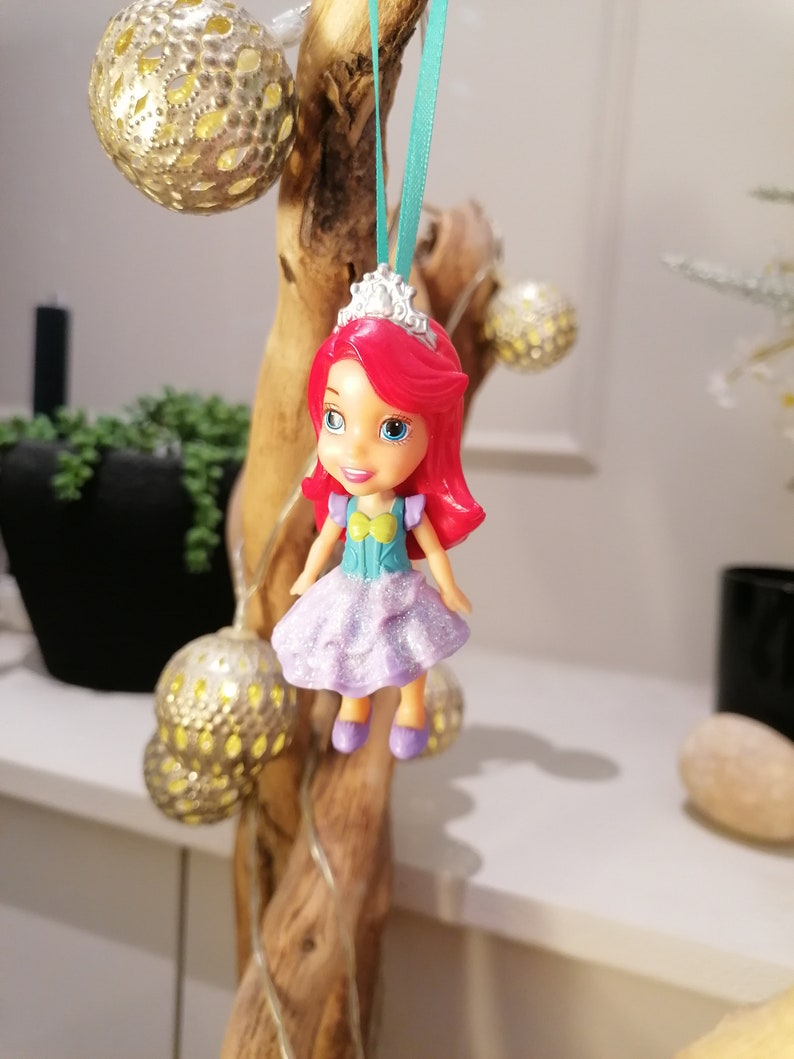 Peut inclure: Une petite poup&eacute;e en plastique aux cheveux roux, v&ecirc;tue d'une robe violette &agrave; paillettes, d'un haut bleu et jaune et d'une couronne argent&eacute;e. La poup&eacute;e est suspendue &agrave; un ruban turquoise sur une branche.