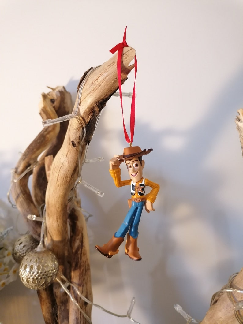 May include: A red ribbon hangs from a branch of driftwood, with a plastic figurine of Woody from Toy Story hanging from the ribbon. Woody is wearing his signature cowboy hat, vest, and jeans. He is holding his right hand up to his hat, as if he is tipping it.