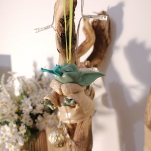 May include: A green and brown Yoda ornament hanging from a branch with a green ribbon. The ornament is made of plastic and has a detailed face.