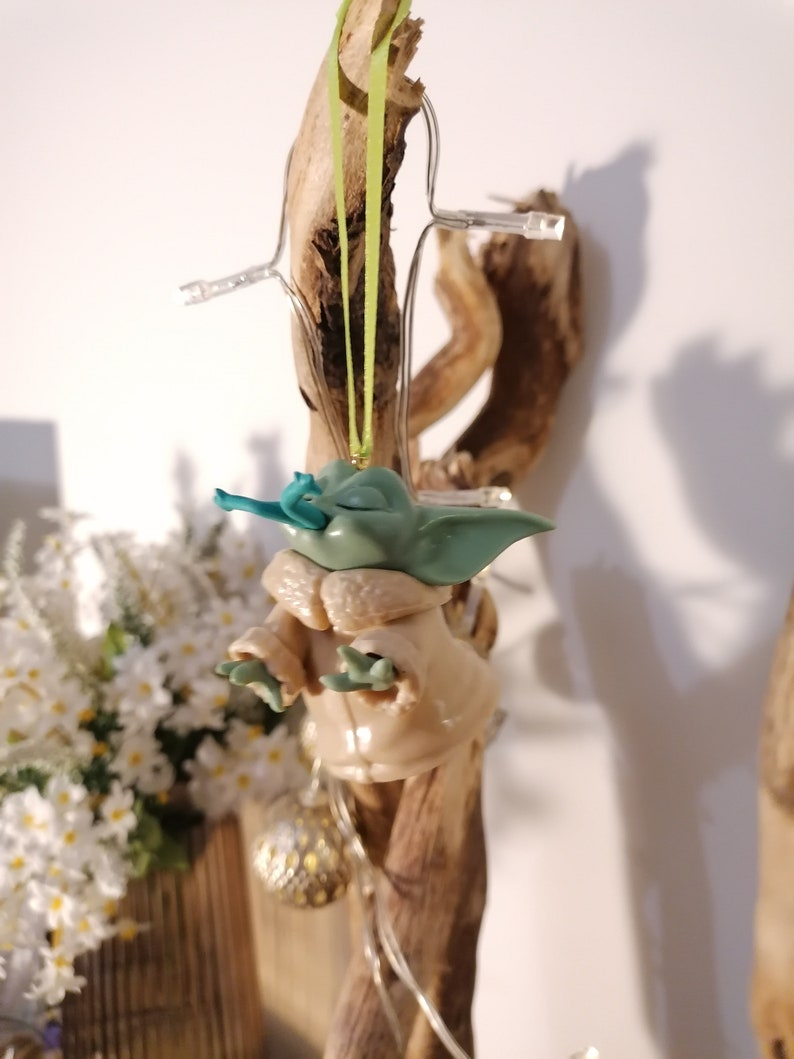 May include: A green and brown Yoda ornament hanging from a branch with a green ribbon. The ornament is made of plastic and has a detailed face.