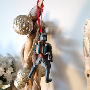 May include: A blue and brown Star Wars character ornament hanging from a red ribbon. The ornament is attached to a piece of driftwood with silver ornaments.