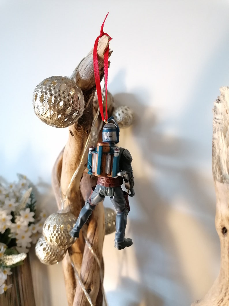 May include: A blue and brown Star Wars character ornament hanging from a red ribbon. The ornament is attached to a piece of driftwood with silver ornaments.
