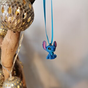May include: A blue Stitch character charm hanging from a turquoise ribbon. The charm is a small, detailed figurine of the popular Disney character.