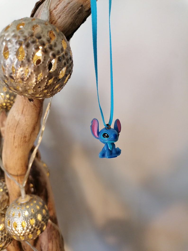 May include: A blue Stitch character charm hanging from a turquoise ribbon. The charm is a small, detailed figurine of the popular Disney character.