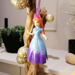 K&ouml;nnte beinhalten: Eine Plastikpuppe mit roten Haaren und einer rosa Krone, die ein lila-blaues Kleid mit Schneeflockenmustern tr&auml;gt. Die Puppe h&auml;ngt an einem lila Band.